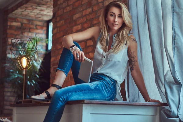 Blogueira feminina loira na moda segura um tablet enquanto se senta em uma mesa contra uma parede de tijolo em um estúdio com um interior de sótão . — Fotografia de Stock