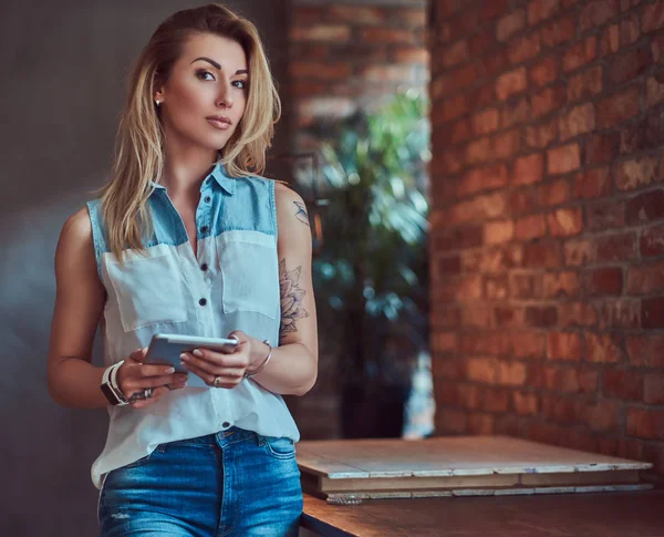 Krásná blondýna ženské blogger pojme tablet přitom stát na stole proti cihlové zdi ve studiu se interiér podkroví. — Stock fotografie