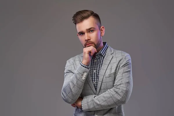 Nadenkend knappe stijlvolle man in een elegante grijze jas, poserend met hand op kin. — Stockfoto