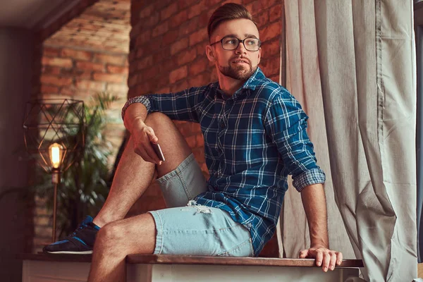 Knappe stijlvolle hipster houdt een smartphone terwijl zittend op een tafel in een kamer met een loft interieur. — Stockfoto
