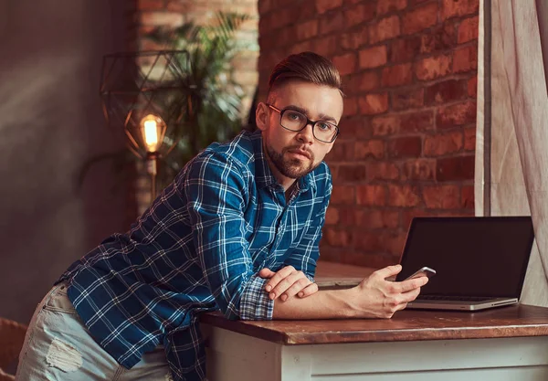 Estudiante guapo sostiene un teléfono inteligente mientras se apoya en una mesa en una habitación con un interior loft . — Foto de Stock