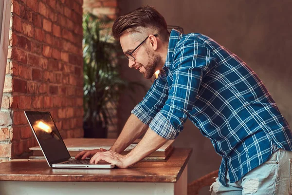 Knappe student in een flanellen shirt werken op een laptop-computer in een kamer met een loft interieur. — Stockfoto