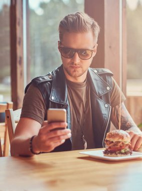 Yenilikçi bir şık saç ve sakal ile yol kenarına yerleştirilen bir kafede öğle yemeği sırasında bir smartphone kullanarak bir masada oturur.