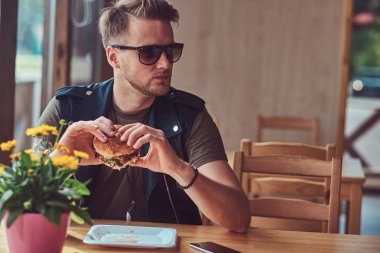 Bir hamburger yeme bir şık saç ve sakal ile yakışıklı hipster yol kenarına yerleştirilen bir kafede yemek yemeye karar verdim bir masada oturur.