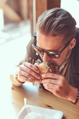 Bir şık saç ve sakal aç hipster adamla portresi bir hamburger yeme yol kenarına yerleştirilen bir kafede yemek yemeye karar verdim bir masada oturur.