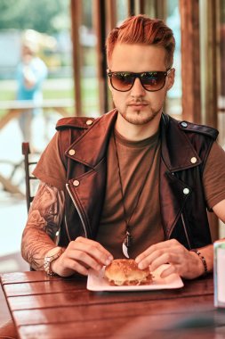 Yakışıklı şık hipster adam portresi bir hamburger ve soda sipariş bir masada oturuyor, yol kenarındaki café'de akşam.
