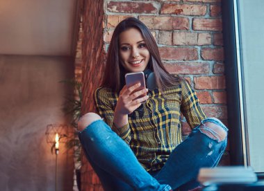 Neşeli esmer öğrenci kız rahat kıyafetler bir pencere pervazına çatı iç ile bir odada otururken bir akıllı telefon kullanırken müzik dinleme.
