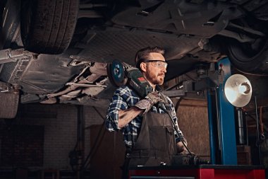 Üniforma ve koruyucu gözlük mekanik onarım garajda araba kaldırma altında duran bir açı değirmeni tutar.