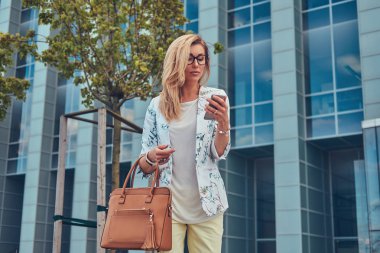 Şık giysiler ve gözlük bir çanta ile moda kadın blogger tutan bir gökdelen karşı duran bir akıllı telefon.