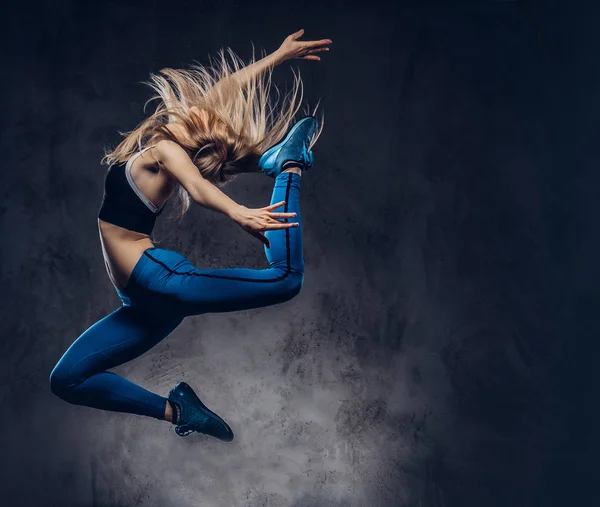 Jonge blonde ballerina in sportkleding dansen en springt in een studio. Geïsoleerd op een donkere achtergrond. — Stockfoto