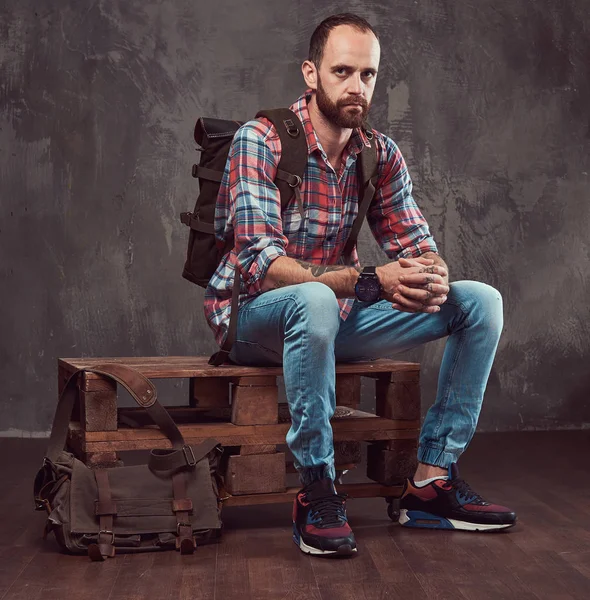 Портрет красивого татуированного путешественника в фланелевой рубашке с рюкзаком, сидящего на скамейке в студии . — стоковое фото