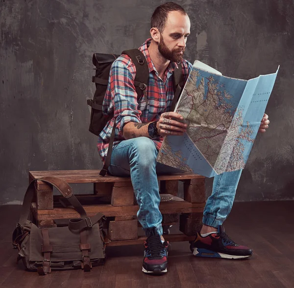 Portrét pohledný Tetovaný cestovatel v flanelovou košili s batohem, obsahuje mapu, sedí na lavičce v ateliéru. — Stock fotografie