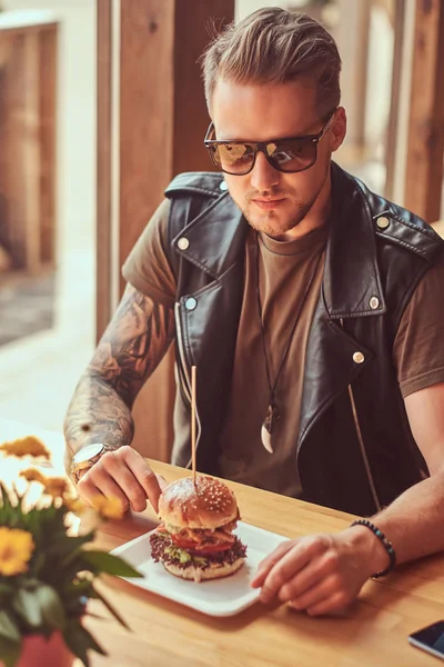 Handsome hipster with a stylish haircut and beard sits at a table, decided to dine at a roadside cafe, eating a hamburger. — Stock Photo, Image