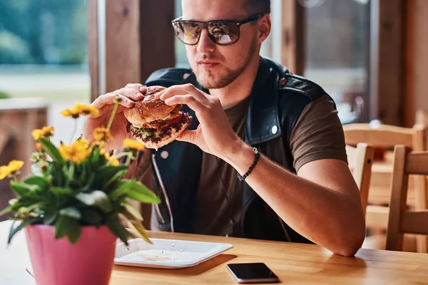 Bonito hipster com um corte de cabelo elegante e barba senta-se a uma mesa, decidiu jantar em um café à beira da estrada, comer um hambúrguer . — Fotografia de Stock