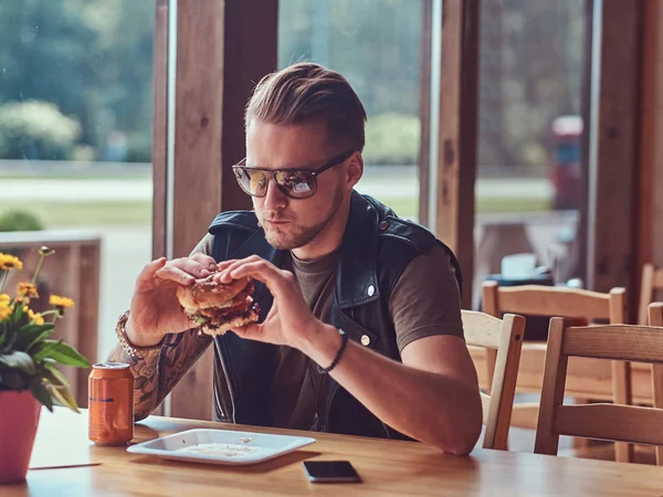 Handsome hipster with a stylish haircut and beard sits at a table, decided to dine at a roadside cafe, eating a hamburger. — Stock Photo, Image
