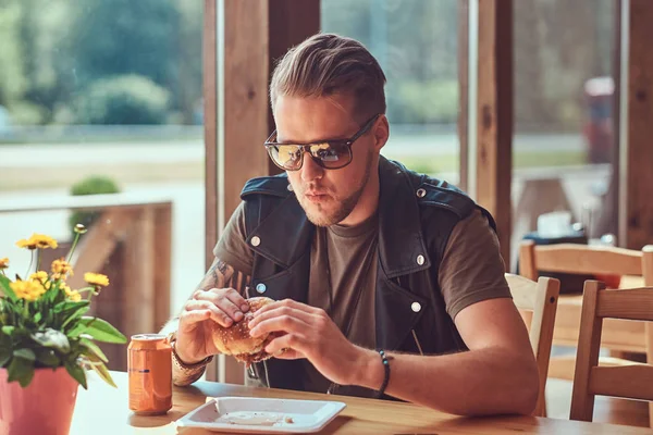 Bonito hipster com um corte de cabelo elegante e barba senta-se a uma mesa, decidiu jantar em um café à beira da estrada, comer um hambúrguer . — Fotografia de Stock