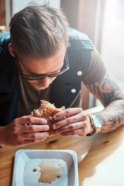 Portrait d'un hipster affamé avec une coupe de cheveux et une barbe élégantes assis à une table, a décidé de dîner dans un café en bord de route, en mangeant un hamburger . — Photo