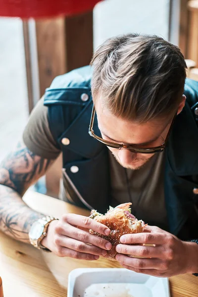 Portrait d'un hipster affamé avec une coupe de cheveux et une barbe élégantes assis à une table, a décidé de dîner dans un café en bord de route, en mangeant un hamburger . — Photo