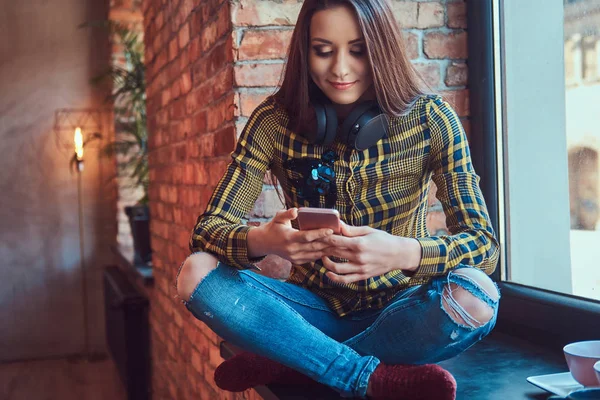 Νεαρός φοιτητής μελαχρινή κοπέλα σε casual ρούχα, ακούγοντας μουσική ενώ χρησιμοποιώντας ένα smartphone, ενώ κάθεται σε ένα περβάζι σε ένα δωμάτιο με εσωτερικό πατάρι. — Φωτογραφία Αρχείου