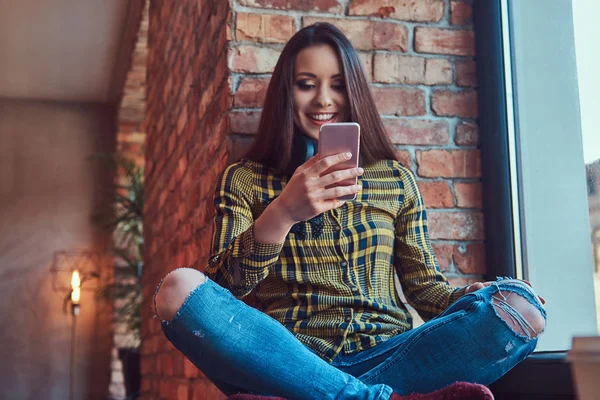 Φοιτητής εύθυμος μελαχρινή κοπέλα σε casual ρούχα, ακούγοντας μουσική ενώ χρησιμοποιώντας ένα smartphone, ενώ κάθεται σε ένα περβάζι σε ένα δωμάτιο με εσωτερικό πατάρι. — Φωτογραφία Αρχείου