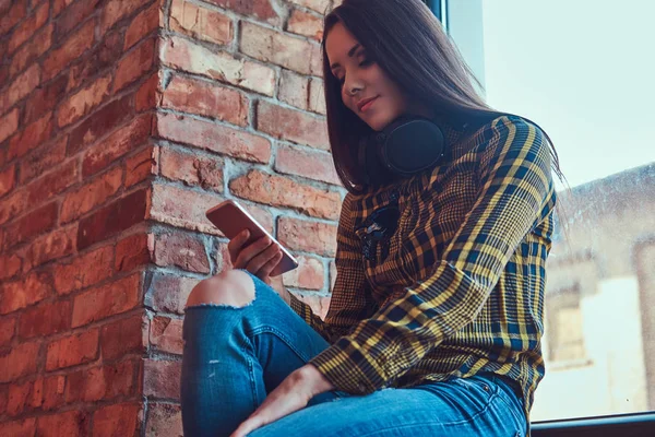 캐주얼 옷 로프트 인테리어와 함께 룸에서 창틀에 앉아있는 동안 스마트폰 사용 하는 동안 음악을 듣고 젊은 갈색 머리 학생 여자. — 스톡 사진