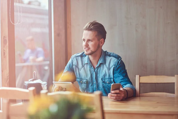 Hipster τύπος με ένα μοντέρνο κούρεμα και γενειάδα κάθεται σε ένα τραπέζι σε μια άκρη του δρόμου café, κατέχει ένα tablet και το smartphone. — Φωτογραφία Αρχείου