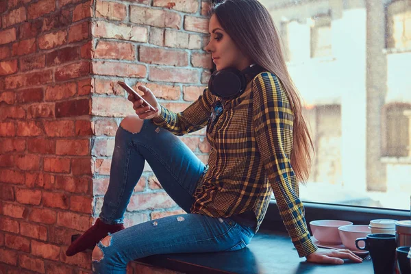 Giovane studentessa bruna in abiti casual ascoltare musica mentre si utilizza uno smartphone mentre seduto su un davanzale di una finestra in una stanza con interni soppalco . — Foto Stock