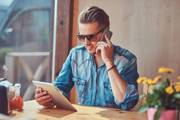Csípő srác egy stílusos hajvágás és szakáll ül egy asztalnál, egy út menti kávézó, beszél a telefonon, és tart egy tabletta számítógép. — Stock Fotó