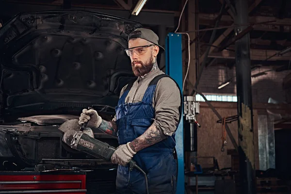 Brutal mecánico de automóviles en un uniforme y gafas de seguridad que trabajan con una amoladora angular mientras está de pie contra un coche roto en el garaje de reparación . — Foto de Stock