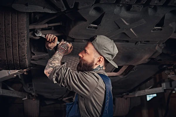 Barbudo mecánico de automóviles en una reparación uniforme de la suspensión de los coches con una llave inglesa, mientras que de pie debajo del coche de elevación en el garaje de reparación . — Foto de Stock