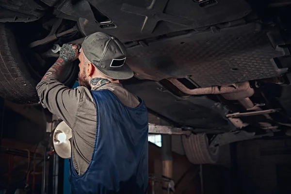 Mécanicien auto barbu dans un uniforme réparer la suspension des voitures avec une clé tout en se tenant debout sous la voiture de levage dans le garage de réparation . — Photo