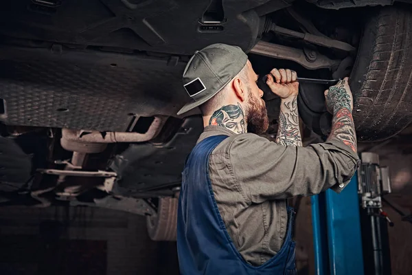 Bearded auto mechanic in a uniform repair the cars suspension with a wrench while standing under lifting car in repair garage.