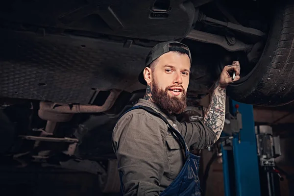 Mécanicien auto barbu dans un uniforme réparer la suspension des voitures avec une clé tout en se tenant debout sous la voiture de levage dans le garage de réparation . — Photo