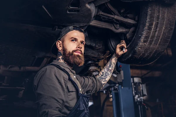 Brodaty auto mechanik w mundurze naprawy zawieszenia samochodów z kluczem dynamometrycznym, stojąc pod podnoszenia samochodu w garażu napraw. — Zdjęcie stockowe