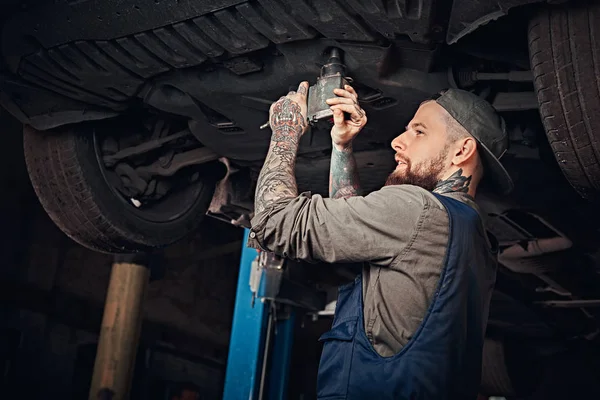 Auto mechanic in a uniform, repair the car with a screwdriver while standing under lifting car in the repair garage.
