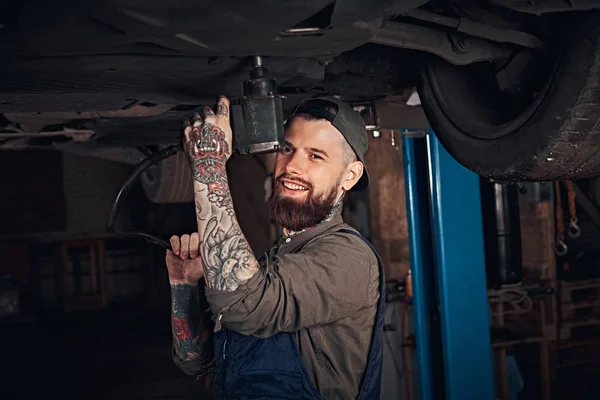 Auto mechanik w jednolity, naprawić samochód śrubokrętem stojąc pod podnoszenia samochodu w garażu napraw. — Zdjęcie stockowe
