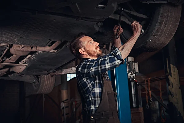 Brodaty auto mechanik w mundurze naprawy zawieszenia samochodów z kluczem dynamometrycznym, stojąc pod podnoszenia samochodu w garażu napraw. — Zdjęcie stockowe