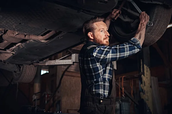 Mécanicien auto barbu dans un uniforme réparer la suspension des voitures avec une clé tout en se tenant debout sous la voiture de levage dans le garage de réparation . — Photo