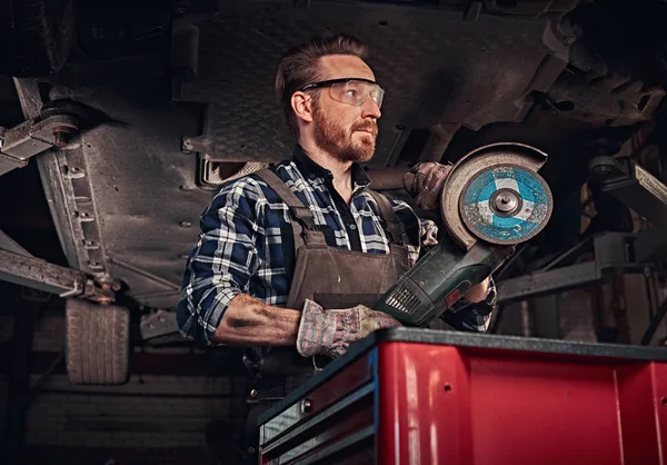 Mécanicien dans un uniforme et lunettes de sécurité tient une meuleuse d'angle tout en se tenant sous la voiture de levage dans un garage de réparation . — Photo