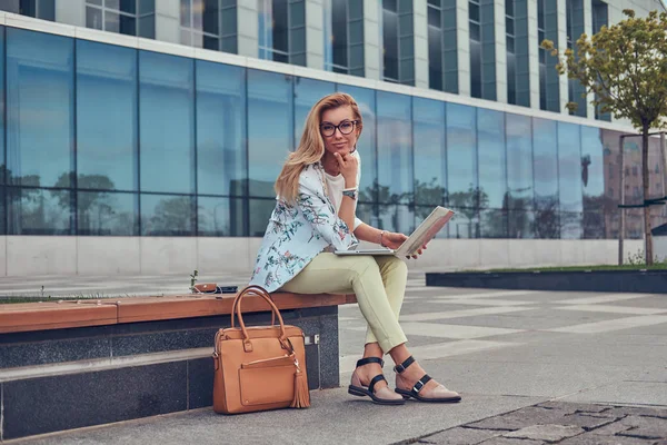 Stylische Modebloggerin, die ihren Laptop zur Arbeit benutzt, während sie draußen auf einer Bank vor einem Wolkenkratzer sitzt. — Stockfoto