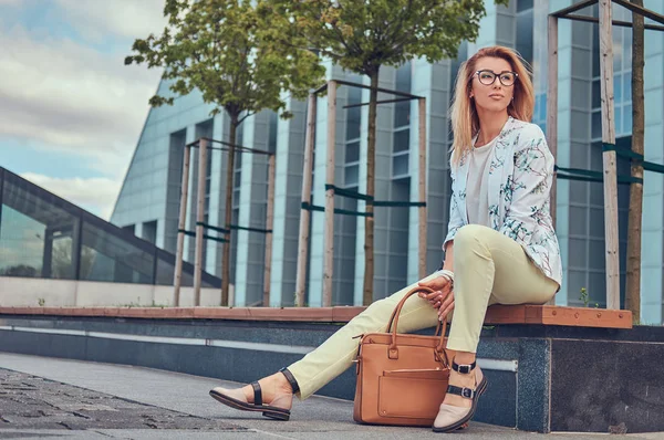 Vackra fashionabla kvinna i snygga kläder och glasögon med en handväska, sitter på en bänk mot en skyskrapa. — Stockfoto