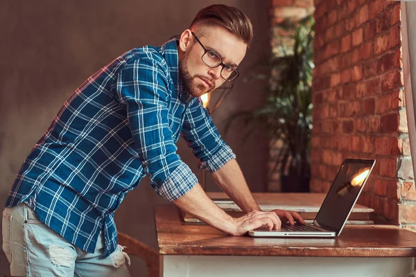 Handsome student in a flannel shirt working on a laptop computer in a room with a loft interior. — Stock Photo, Image