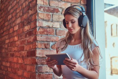 Genç Sarışın kadın müzik dinlemek ve bir pencere pervazına bir çatı katı iç ile bir odada otururken dijital tablet kullanma.