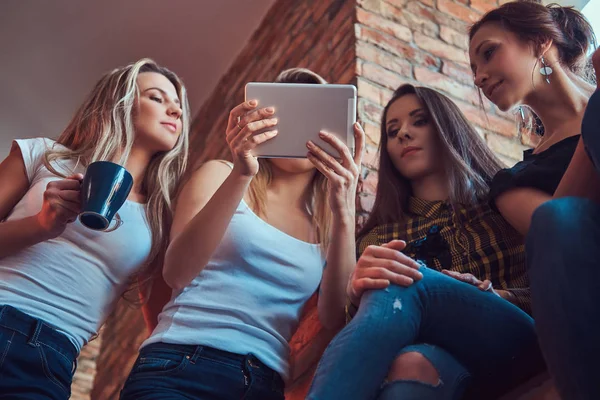 Group of female friends in casual clothes discussing while looking something on a digital tablet in a room with a loft interior.