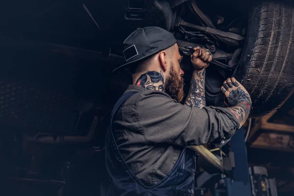 Sakallı otomatik mekanik bir üniformalı tamir onarım garajda araba kaldırma altında duran bir anahtar ile araçların süspansiyon. 