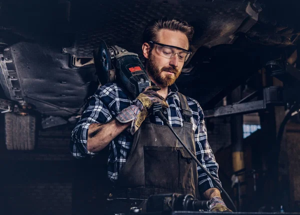 Auto mechanic in a uniform and safety glasses holds an angle grinder while standing under lifting car in a repair garage.