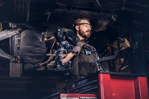 Auto mechanic in a uniform and safety glasses holds an angle grinder while standing under lifting car in a repair garage.