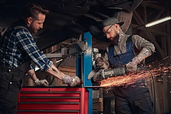 Two bearded auto mechanics in a uniform and safety glasses working with an angle grinder while standing under lifting car in a repair garage.