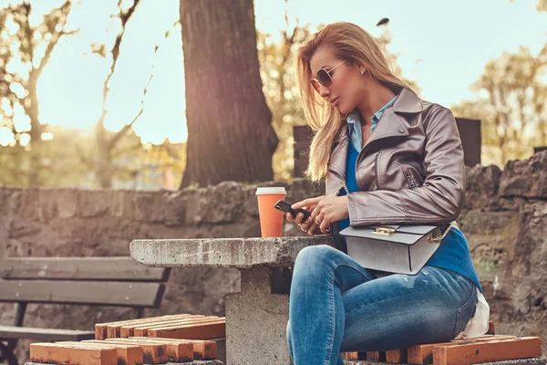 Trendy bionda blogger femminile si rilassa all'aperto, seduto sulla panchina nel parco cittadino contro una luce solare brillante . — Foto Stock