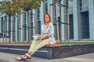 Bir gökdelen karşı parkta bir bankta oturmuş bir kitapla eğitim modern giysiler, çekici sarışın kadın.
